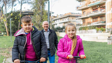 Two children ride scooters while their grandfather walks behind them.